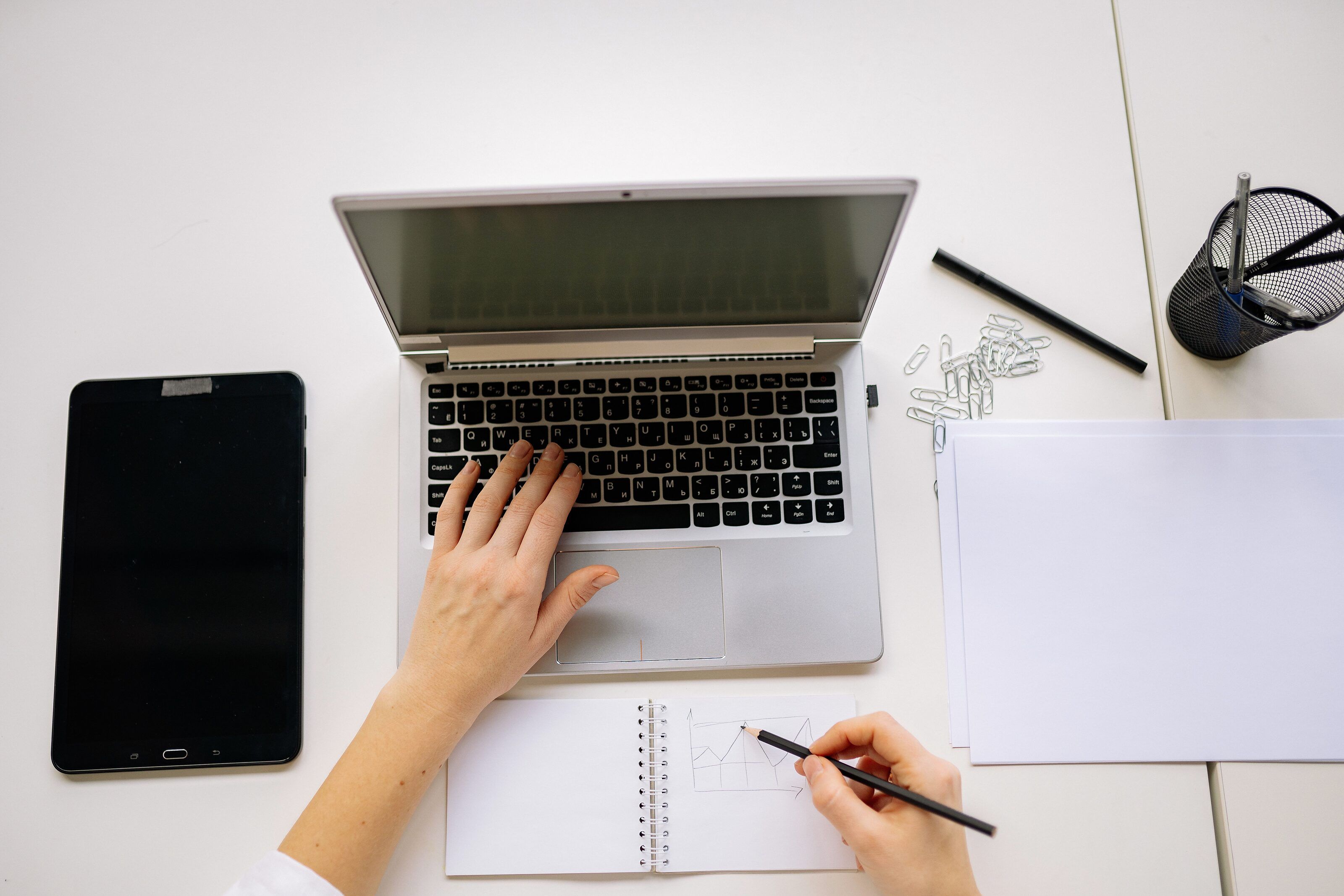 White desk from above: one hand types on a laptop while the other sketches a chart in a notebook, beside a tablet and blank paper.
