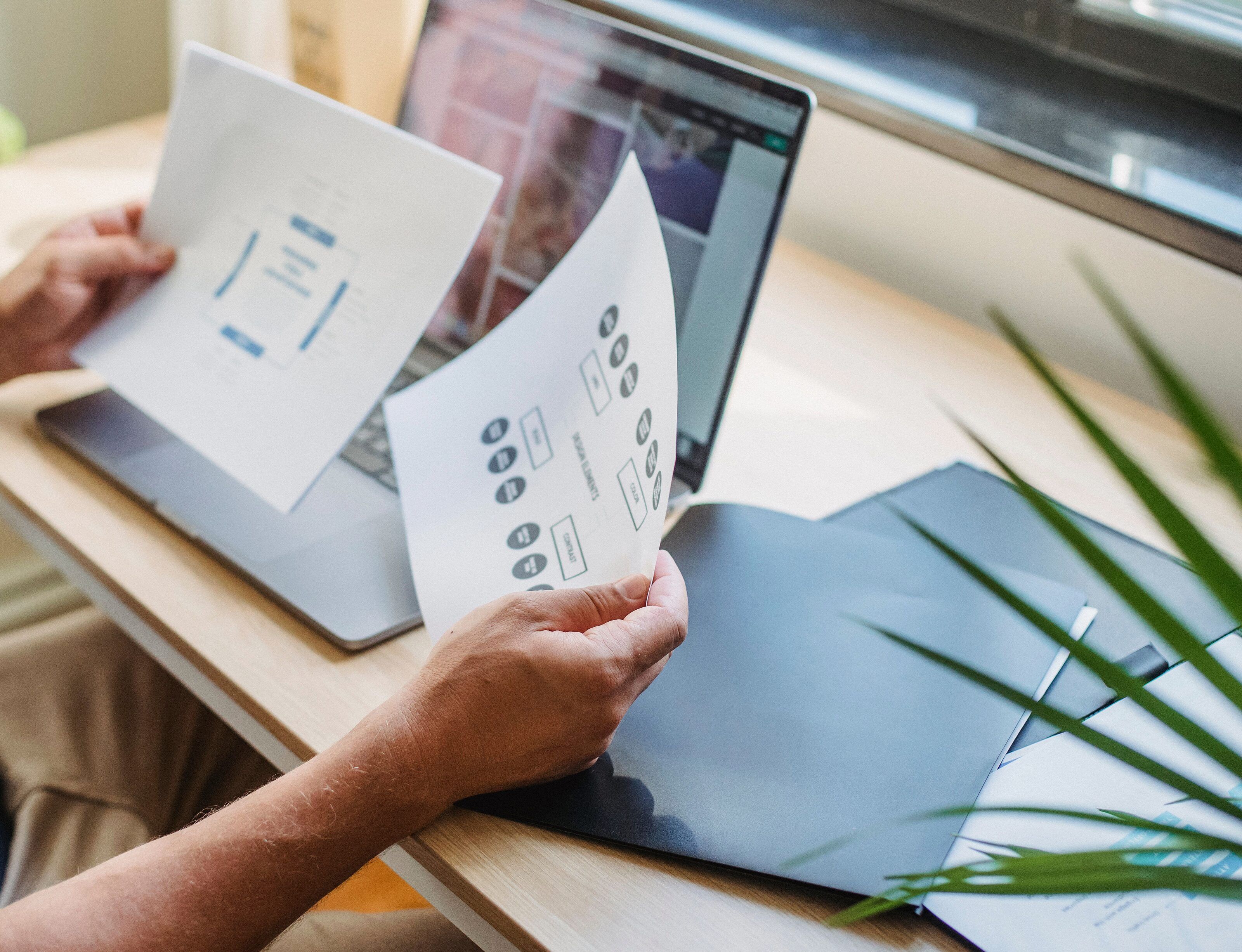 Hands holding two printed interface layouts in front of an open laptop on a wooden desk.