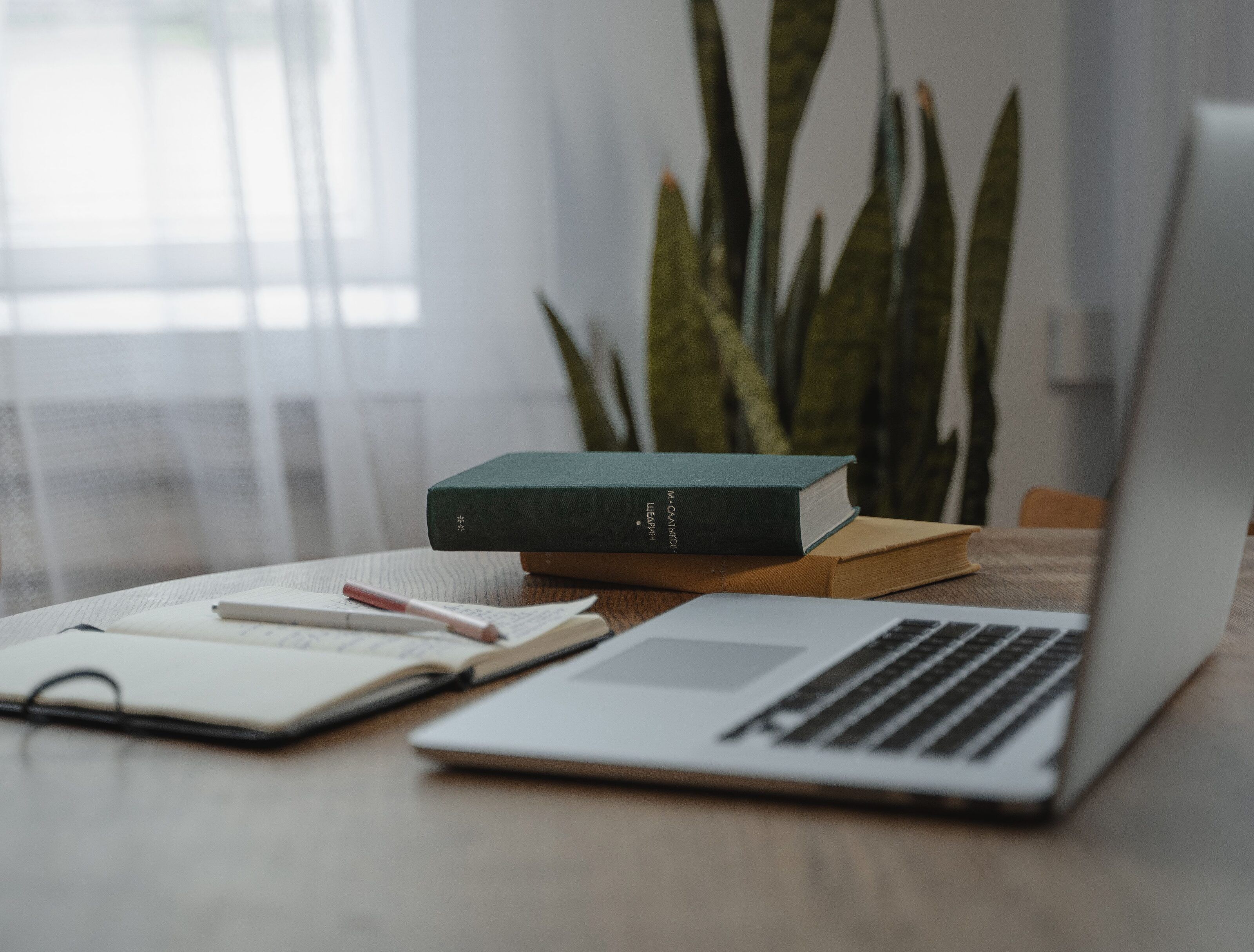 Open notebook with two pens, two hardback books and a laptop on a round wooden table by a curtained window.