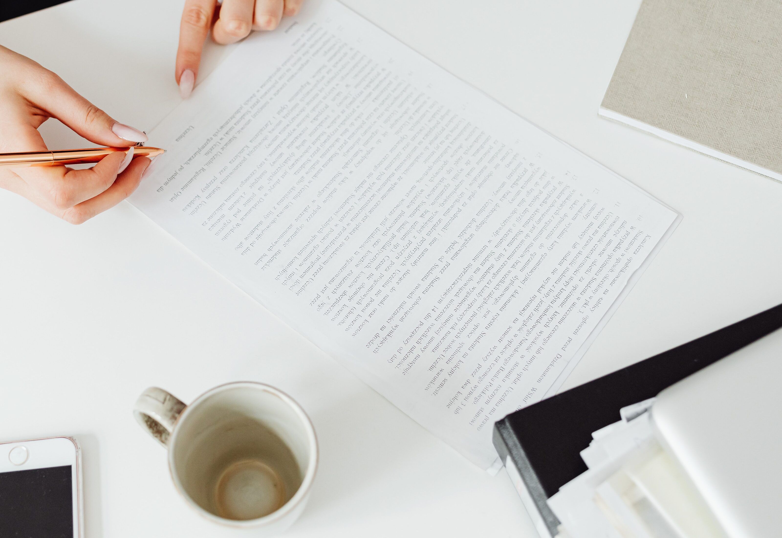 One hand holds a pen over a printed document on a white desk while the other points at the text, with a mug, a notebook and the corner of a phone nearby.