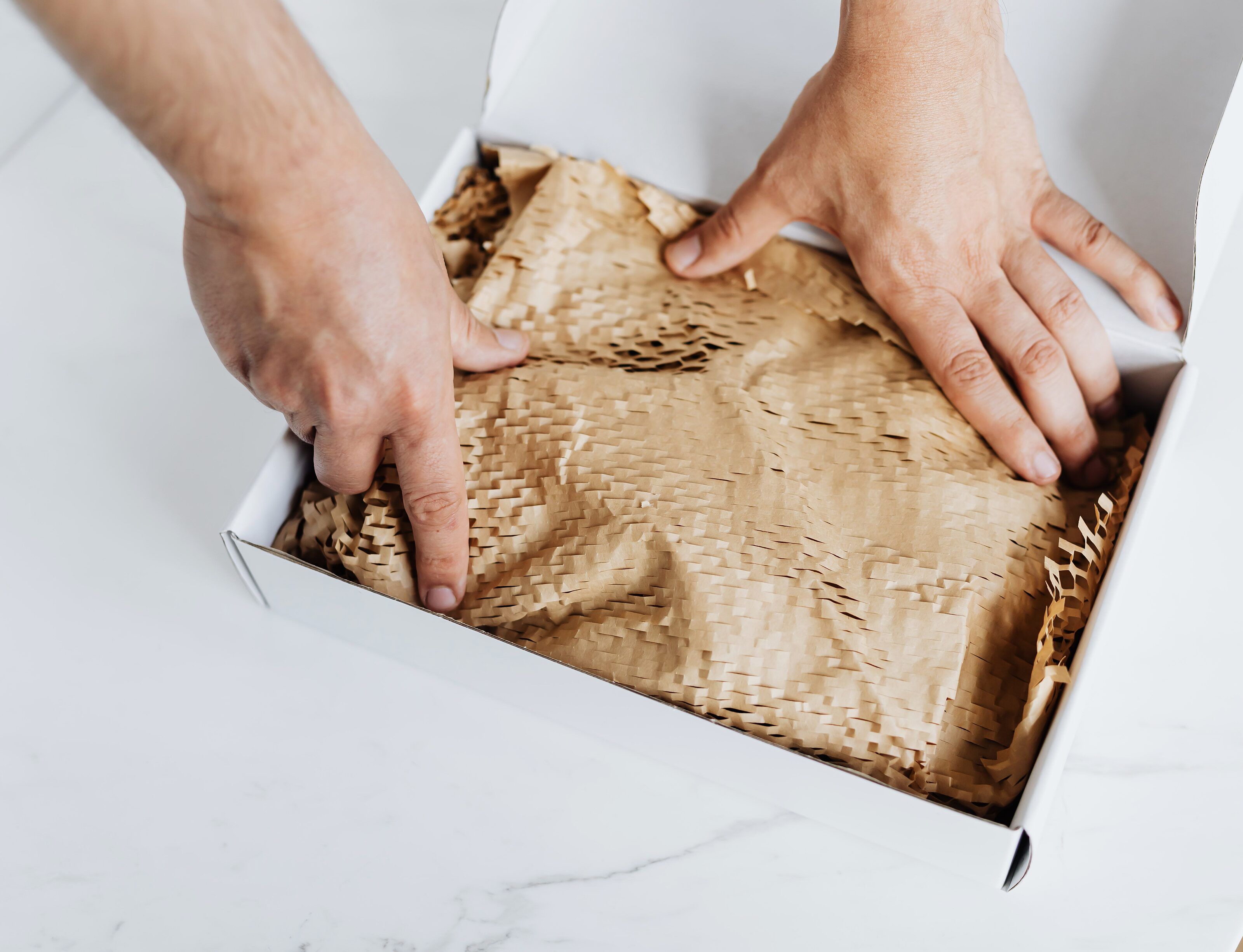 Two hands pressing kraft honeycomb packing paper into an open white shipping box on a white marble table.