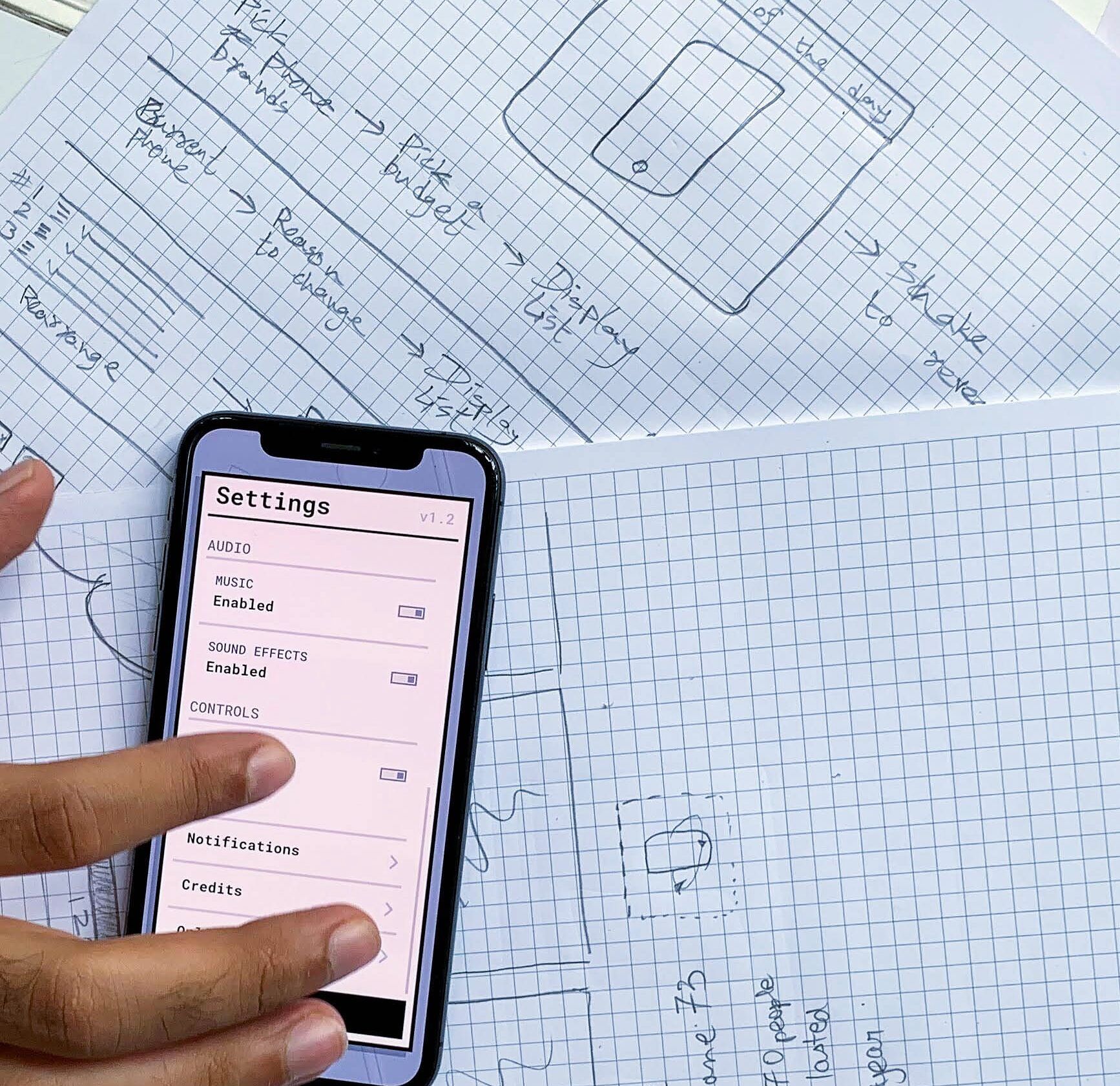 Hand using a settings screen on a smartphone laid over hand-drawn app wireframes on grid paper.